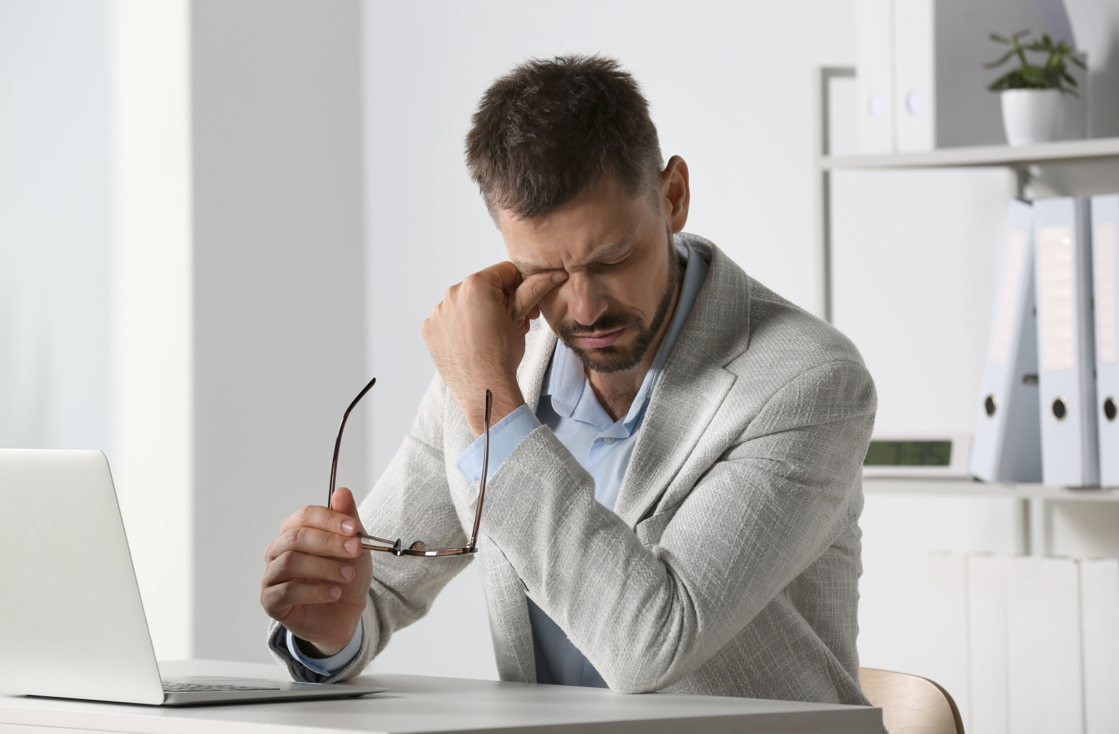 A man sitting at a desk in an office with his laptop and holding his glasses in his right hand as he rubs his eyes.