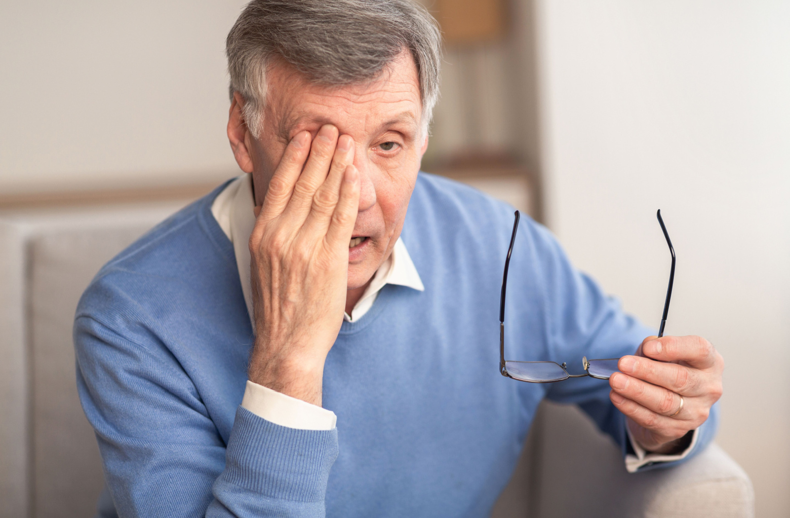 A senior man sitting on a couch holding his glasses in his left hand as he rubs his right eye with his right hand.