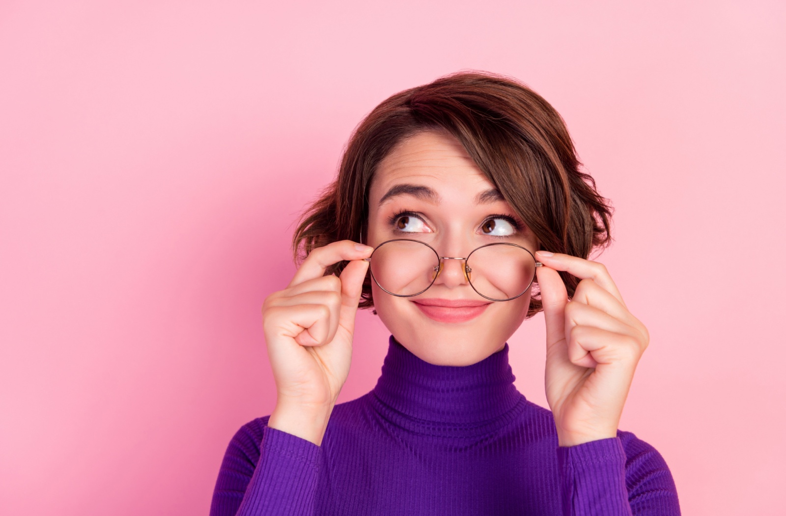 A young woman holding spectacles above her nose with both hands and looking towards her extreme left.