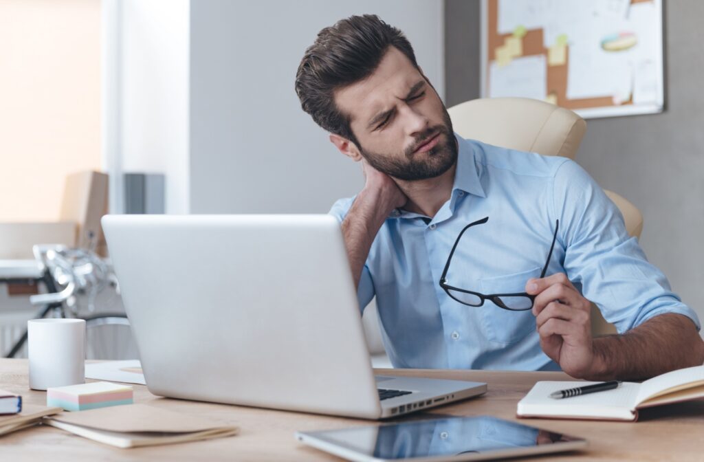 A man holding the back of his neck in pain with one hand and a pair of glasses on the other hand while sitting in front of a computer screen.