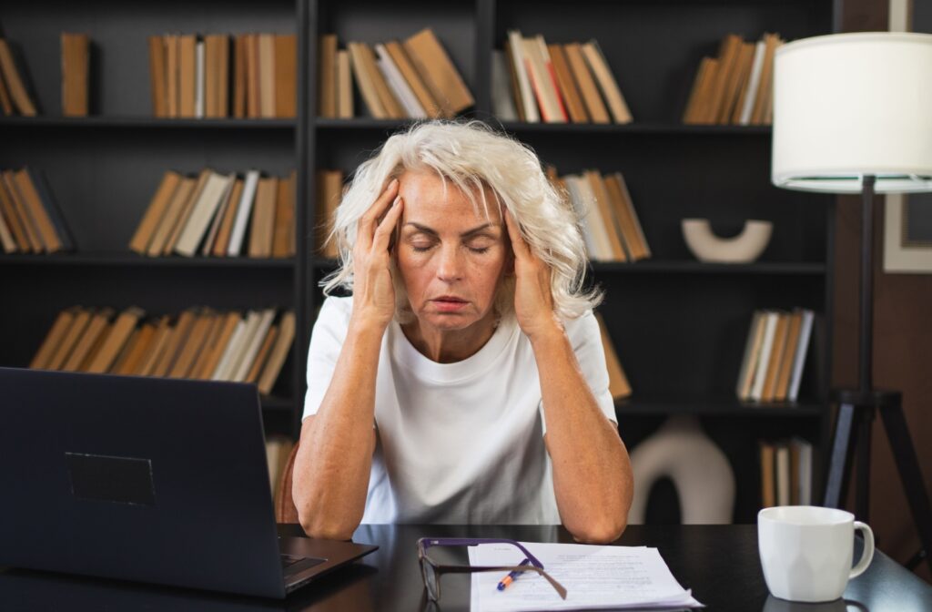 A mature woman suffering from a headache. She is holding the sides of her head, with her eyes closed and a pained expression on her face.