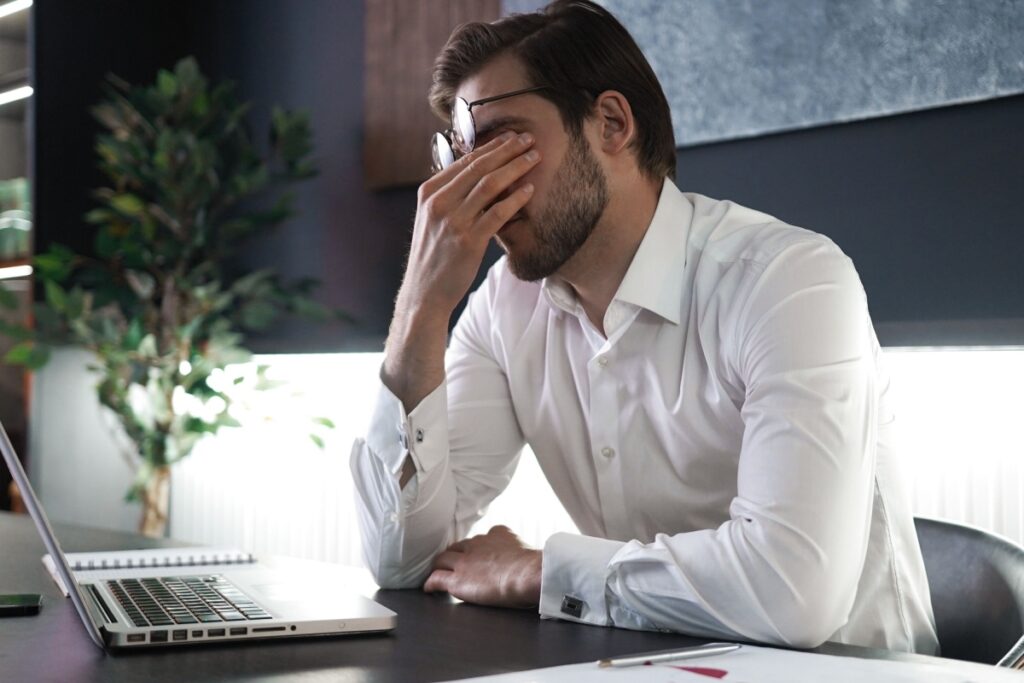Man in white shirt rubbing eyes with hand while sitting at desk with laptop showing symptoms of digital eye strain and fatigue in modern office setting.